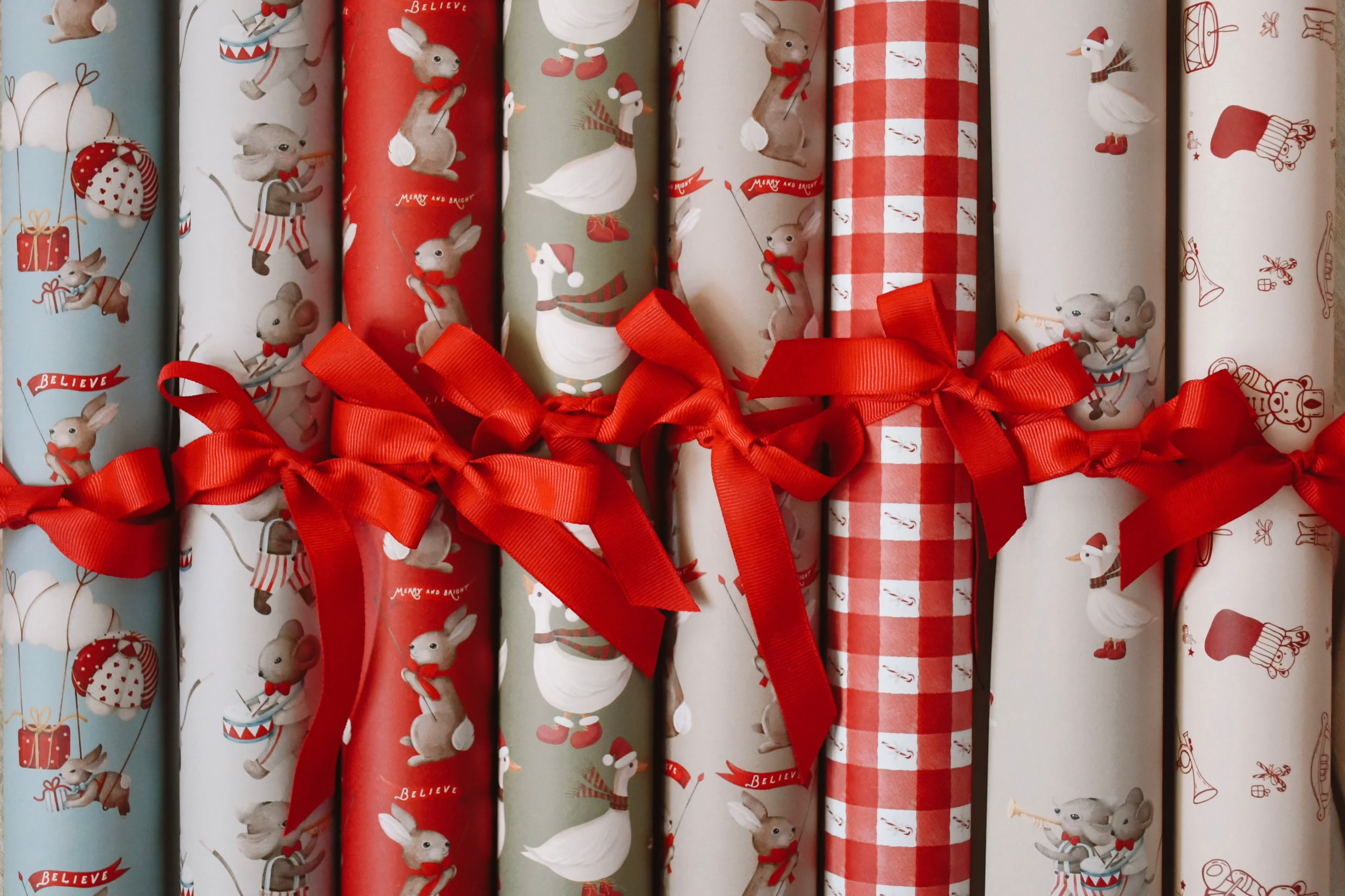 A set of rolled Christmas-themed wrapping paper tied together with red ribbon bows. The patterns include gray bunnies with red scarves and hats on a light blue background, gray bunnies on a white background, red and white ducks, red and white gingham, and white snowmen on a light gray background.